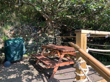 A wooden picnic table next to a green covered barbecue with a stone wall and trees in the background at Windycroft in Barmouth