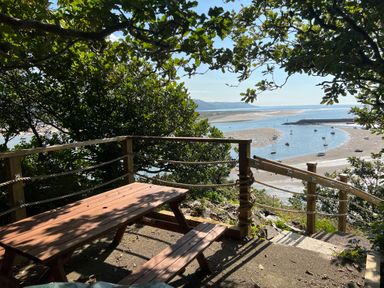 A wooden picnic table and benches on a patio overlooking a river with boats and a sandy shore surrounded by trees at Windycroft in Barmouth