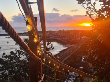 A sunset view over a coastal town with a wooden railing and string lights overlooking rail tracks and the sea at Windycroft in Barmouth