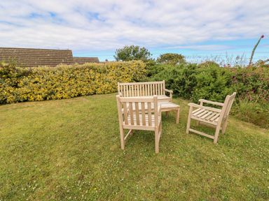 A garden with wooden chairs and a bench on grass surrounded by bushes at Chalfonts Cottage in Trefin near Penparc