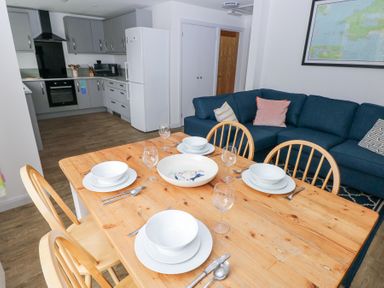 A dining table set with plates bowls and glasses next to a blue sofa and a modern kitchen in the background at Chalfonts Cottage in Trefin near Penparc