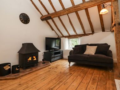 A room with wooden beams a wood stove a television on a cabinet and a black sofa with cushions at 1 White House Cottages Bromsash near Lea