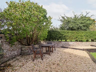 A garden patio with a round wooden table and two wooden chairs surrounded by stone walls and green hedges at 1 White House Cottages Bromsash near Lea
