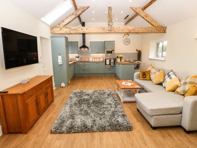 A living room and kitchen area with a gray sofa a wooden coffee table a wooden sideboard a wall-mounted TV and exposed wooden beams at Y Felin The Granary in Caernarfon