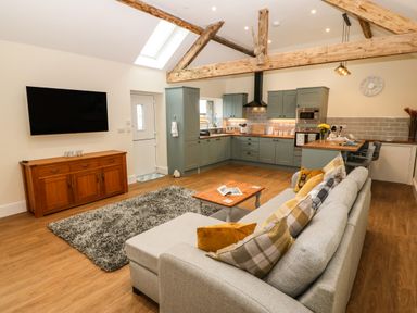 An open plan living room and kitchen with a grey couch pillows wooden beams and a wall mounted TV at Y Felin The Granary in Caernarfon