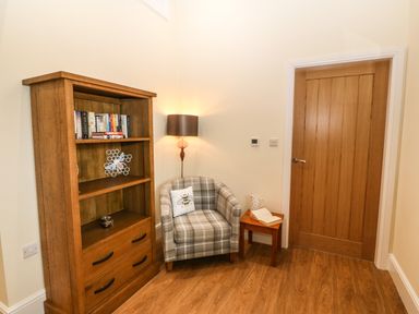 A corner with a wooden bookcase containing books and decor a plaid armchair with a bee pillow a small wooden table with a book and jug a floor lamp and a wooden door at Y Felin The Granary in Caernarfon