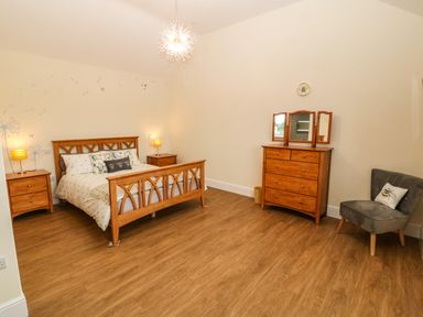 A bedroom with a wooden bed between two nightstands a wooden dresser with a three-part mirror and a gray chair with a pillow at Y Felin The Granary in Caernarfon