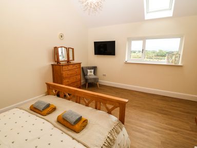 A bedroom with a wooden bed, dresser with mirror, upholstered chair, wall-mounted TV, and window showing an outdoor view at Y Felin The Granary in Caernarfon