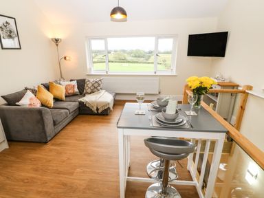 A living room with a gray sofa yellow pillows a dining table set for two and a wall-mounted TV at Ty Gwair The Hayloft in Caernarfon