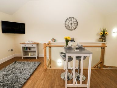 A dining area with a small table set for two and bar stools next to a wooden stair railing and a wall clock at Ty Gwair The Hayloft in Caernarfon