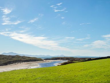 A grassy field with a river flowing towards a sandy beach and hills in the background at 7 Bro Branwen Aberffraw near Rhosneigr