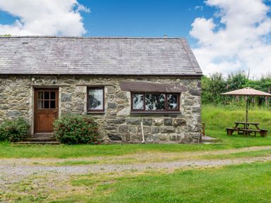 A stone cottage with two windows and a wooden door with bushes in front and a picnic table with an umbrella in the grass at Tryfan Cottage in Rhostryfan near Caernarfon