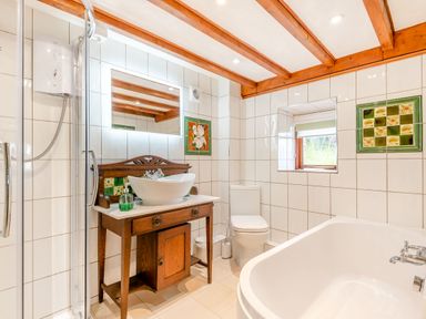 A bathroom with a shower, wooden vanity with sink, toilet, bathtub, window and tiled walls at Tryfan Cottage in Rhostryfan near Caernarfon