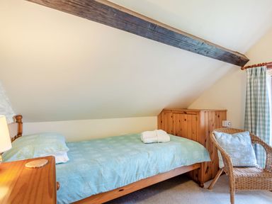 A bedroom with a single bed with light blue bedding beside a wooden table with a lamp and a wicker chair with cushions at Tryfan Cottage in Rhostryfan near Caernarfon