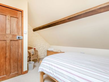 A bedroom with a wooden bed, striped bedding, a wooden door, a wicker chair with a cushion, a small wooden chest, and a sloped ceiling with a wooden beam at Tryfan Cottage in Rhostryfan near Caernarfon
