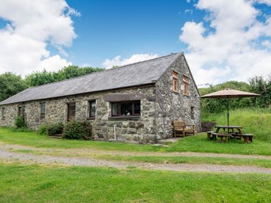 A stone cottage with wooden windows and a slate roof with a wooden bench and a picnic table with an umbrella on grass at Tryfan Cottage in Rhostryfan near Caernarfon