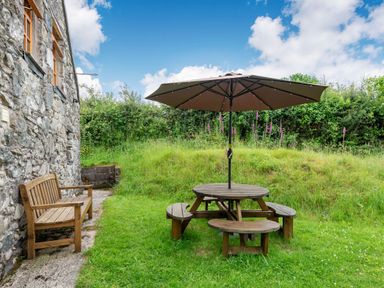 An outdoor garden area with a wooden bench beside a stone wall and a round wooden picnic table with benches and a large umbrella at Tryfan Cottage in Rhostryfan near Caernarfon