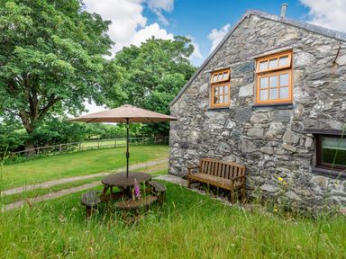 A stone cottage exterior with wooden bench and round table with umbrella in a garden at Tryfan Cottage in Rhostryfan near Caernarfon