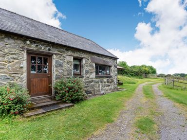A stone cottage with a wooden door and windows next to a gravel and grass driveway at Tryfan Cottage in Rhostryfan near Caernarfon