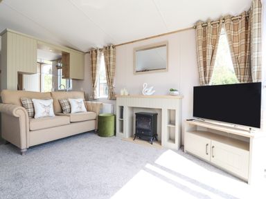A living room with a beige sofa with cushions a small green ottoman a fireplace with decor a wall mirror and a television on a stand at 25 Sherwood in South Lakeland Leisure Village