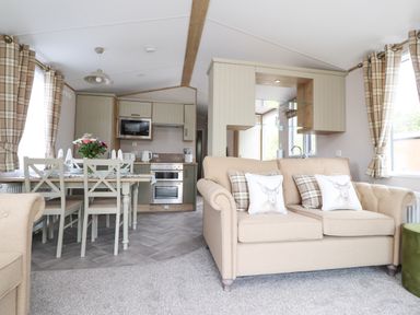 An open living area with beige sofas and plaid cushions beside a dining table with white chairs and a kitchen with appliances at 25 Sherwood in South Lakeland Leisure Village