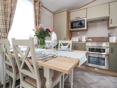 A dining table with chairs set with plates and glasses next to a kitchen with oven microwave and kettle at 25 Sherwood in South Lakeland Leisure Village