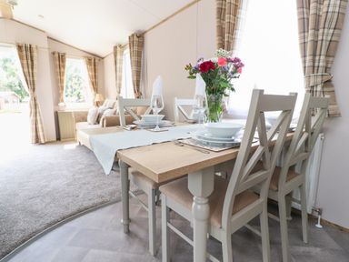 A dining table set with plates bowls glasses and napkins with a vase of flowers in a room with beige chairs and plaid curtains at 25 Sherwood in South Lakeland Leisure Village