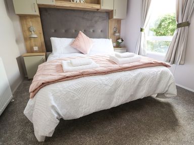 A bedroom with a double bed with white bedding and pink throw and pillows two bedside tables with lamps and a vase of flowers and a window with curtains at 25 Sherwood South Lakeland Leisure Village