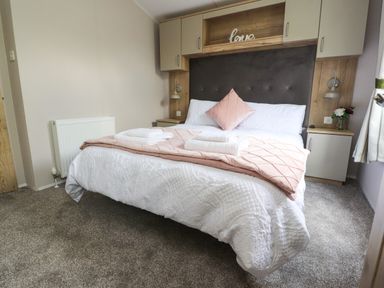 A bedroom with a double bed white bedding pink pillow and towels with storage cabinets above and bedside tables at 25 Sherwood in South Lakeland Leisure Village
