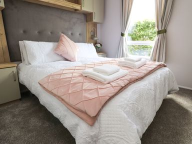 A bedroom with a bed with white bedding and a pink throw with pillows next to a window with curtains at 25 Sherwood in South Lakeland Leisure Village