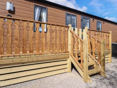 A wooden porch with stairs leading to a door of a brown cabin with windows at 25 Sherwood in South Lakeland Leisure Village