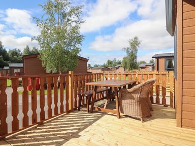 A wooden outdoor deck with a table bench and wicker chair overlooking brown cabins at 25 Sherwood in South Lakeland Leisure Village