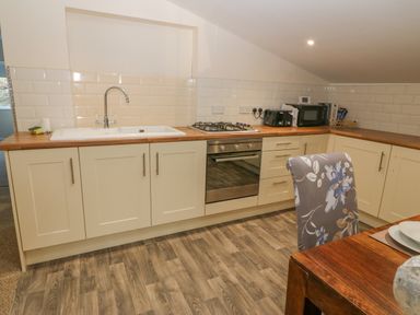 A kitchen with a sink, stove, oven, countertop appliances, cabinets, and a wooden dining table with a floral chair at The Apartment at Plas Yr Eithin in Amlwch near Llanfechell