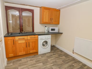 A kitchen area with wooden cabinets a washing machine microwave kettle and a window at The Apartment at Plas Yr Eithin Amlwch near Llanfechell