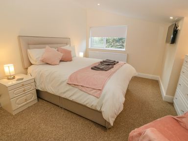 A bedroom with a bed with pillows and folded towels a nightstand with a lamp a window a wall-mounted TV a dresser and a chair at The Apartment at Plas Yr Eithin in Amlwch near Llanfechell