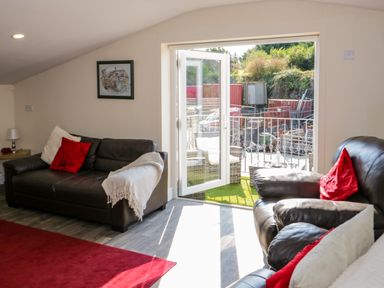 A living room with leather sofas and red cushions by an open glass door leading to an outdoor area at The Apartment at Plas Yr Eithin in Amlwch near Llanfechell
