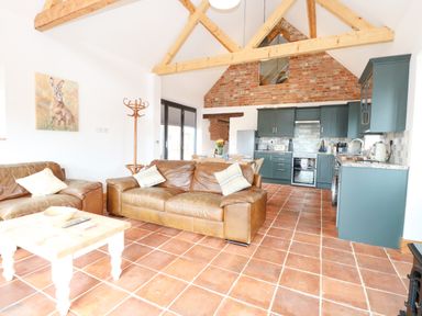 An open living room and kitchen with leather sofas wooden beams and brick wall at Little Barn in Tittleshall