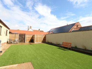 A backyard with green artificial grass a wooden bench a patio table with chairs and a wooden fence at Little Barn in Tittleshall