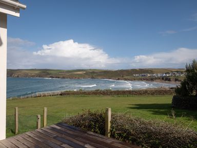 A view of the coastline with waves, grassy area, wooden deck, fence, and houses in the distance at Seascape in Polzeath