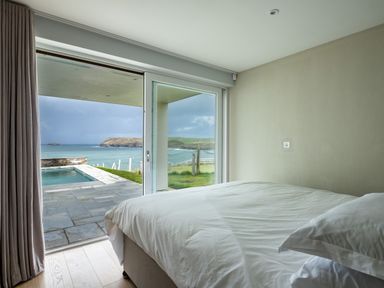 A bedroom with a bed facing glass sliding doors overlooking a patio pool and sea at Seascape in Polzeath