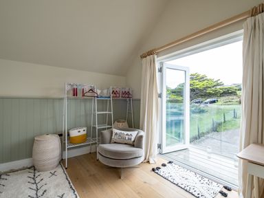 A room with a round armchair a window door open to outside a metal rack with hangers and baskets and a rug on the wooden floor at Seascape Polzeath