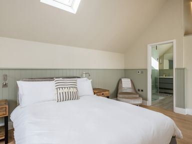 A bedroom with a bed white linens a striped pillow two wooden nightstands a beige chair and a door leading to a bathroom at Seascape in Polzeath