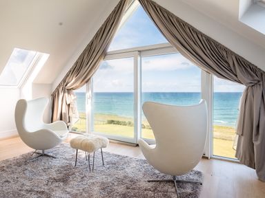 A room with two white modern chairs and a small stool on a rug facing a large window with a sea view at Seascape in Polzeath