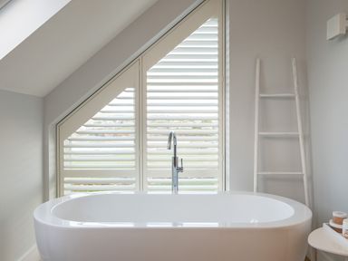A bathroom with a freestanding bathtub under a shuttered triangular window and a ladder rack beside it at Seascape in Polzeath