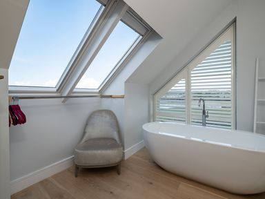 A bathroom with a freestanding bathtub a gray chair and slanted windows at Seascape in Polzeath