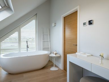 A bathroom with a white freestanding bathtub a slanted window with blinds a wooden door a small round white side table and a white desk with a stool at Seascape in Polzeath