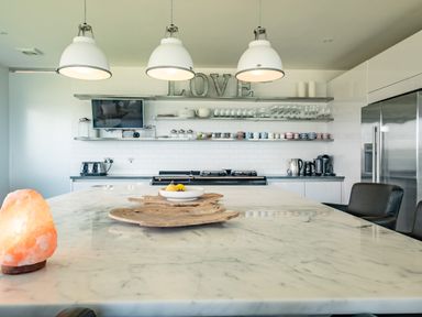 A kitchen with a marble island countertop with wooden trays and a salt lamp and shelves with cups glasses and a tv at Seascape in Polzeath