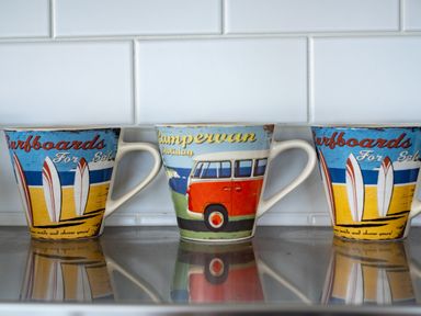 Three mugs with surfboard and campervan designs on a kitchen countertop at Seascape in Polzeath
