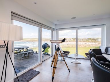 A living room with a telescope, chairs, round table, floor lamp, and large windows overlooking the beach at Seascape in Polzeath