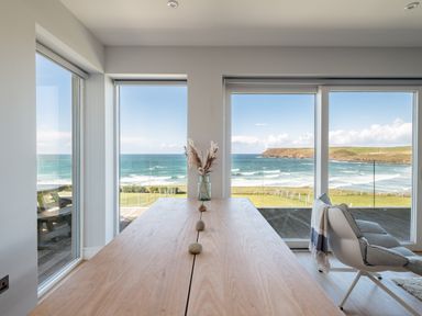 A dining area with a wooden table and a vase with dried plants overlooking the ocean through large windows at Seascape in Polzeath